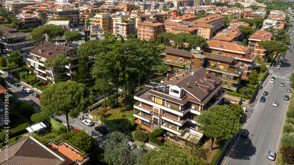 Fototapeta premium High-angle aerial view of a multi-story residential building featuring large balconies, nestled within a leafy neighborhood with tall pine trees, orange roofs, and a street with light car traffic.