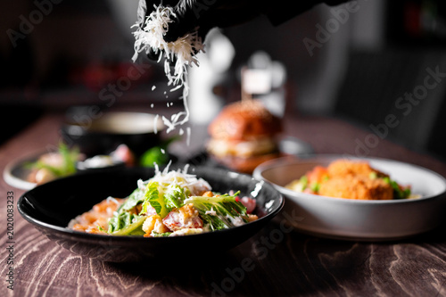 Fotografie A chef sprinkling grated parmesan cheese onto a fresh Caesar salad in a dark restaurant bowl