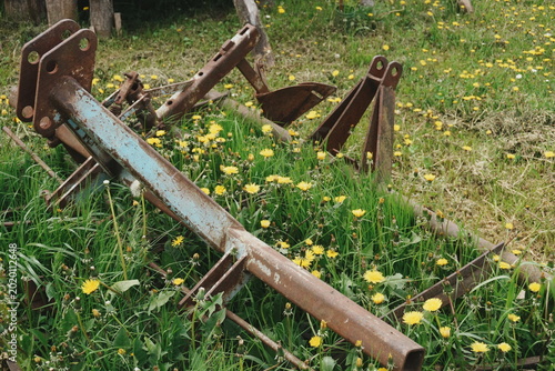Fotografie Weathered Rusty Farm Equipment Plow Parts Resting on Green Grass with Yellow Wil