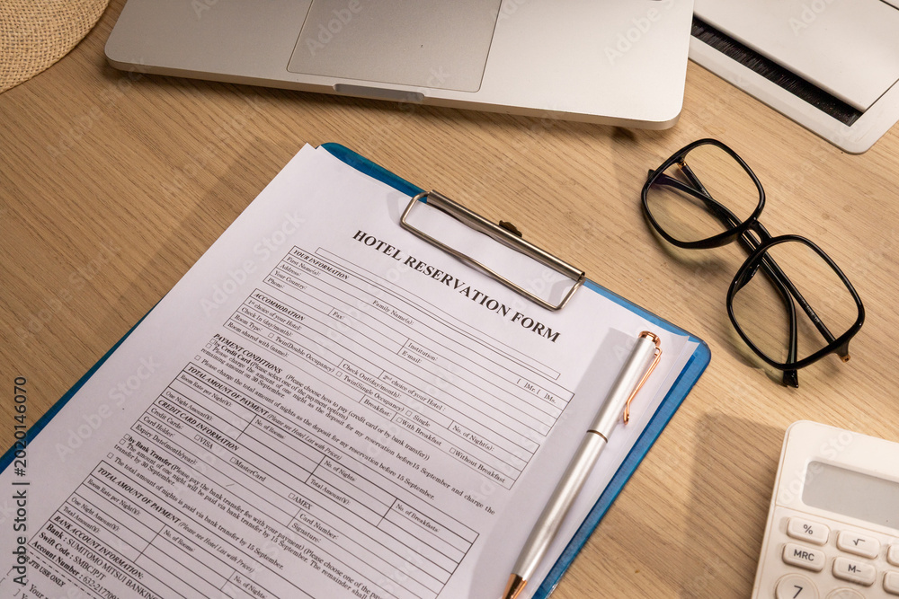 Obraz premium High angle view of a blank Hotel Reservation Form on a blue clipboard with a silver pen and laptop on a wooden desk