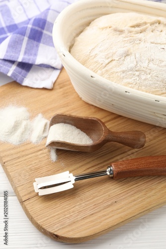 Schilderij op canvas Raw sourdough bread in proofing basket, flour and lame on white wooden table