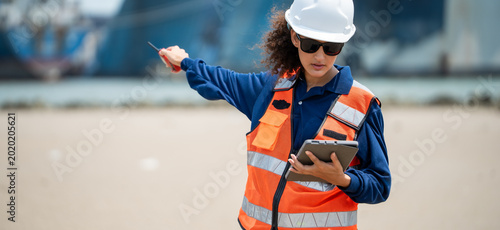 Professional female port supervisor points toward cargo vessels while managing logistics data on a tablet during marine operations at a busy commercial shipping harbor