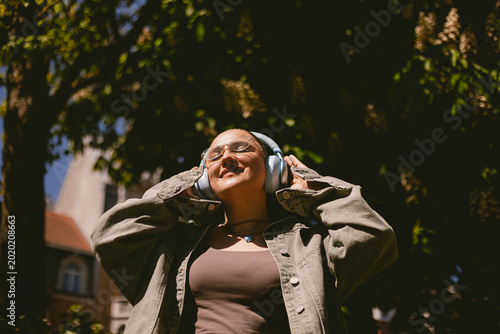 Serene young woman in stylish glasses and denim jacket listening to music in wireless headphones with closed eyes and peaceful smile in sunny park, happy girl enjoying favorite audio in nature