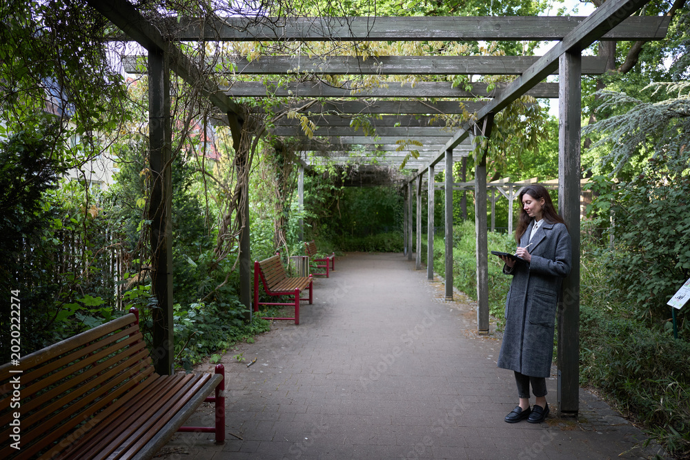 Obraz premium Woman in coat using tablet under a wooden pergola in a lush green garden
