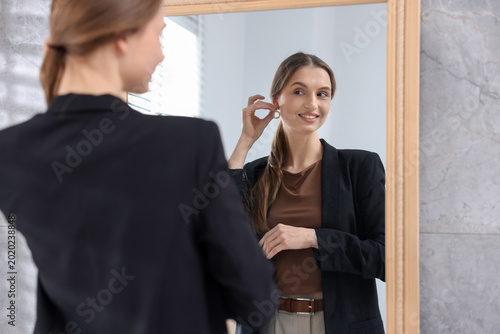 Beautiful young woman wearing stylish earring near mirror indoors  selective focus
