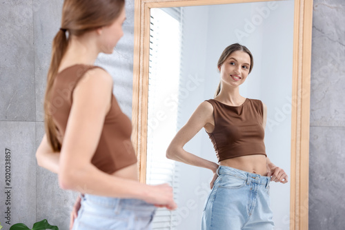 Fotografía Beautiful young woman wearing big jeans looking at herself in mirror indoors, se