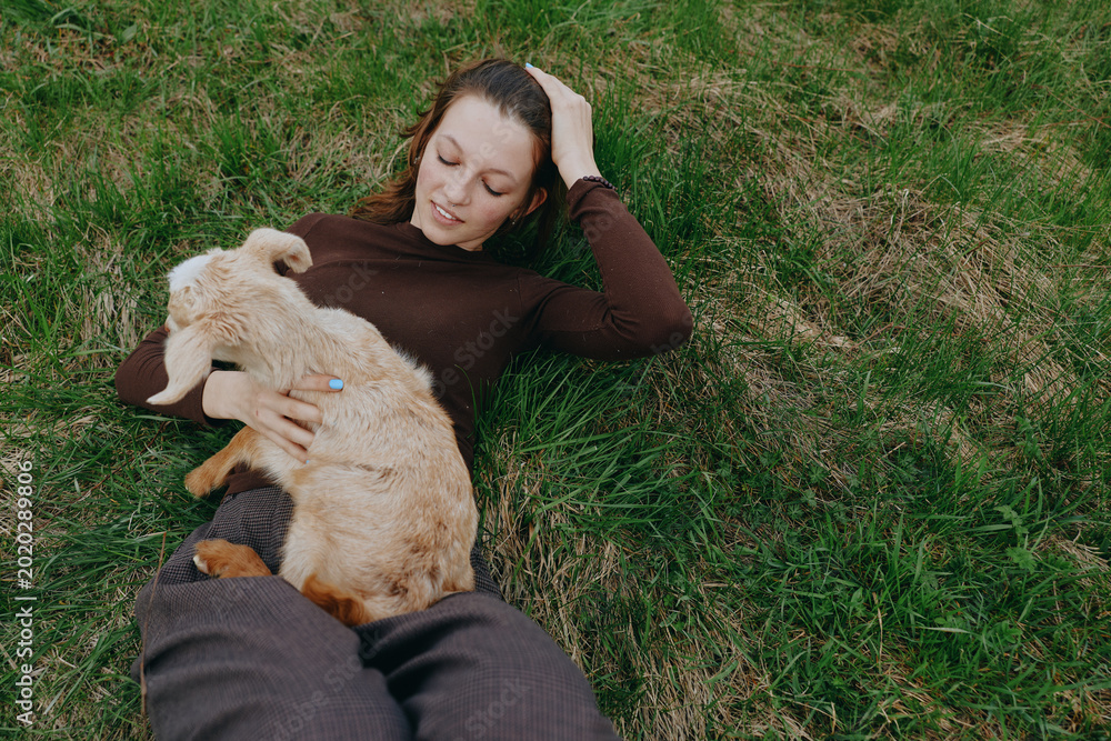 Obraz premium Relaxed woman lies on green grass outdoors, cuddling a young goat kid, sharing a gentle moment of companionship and calm, natural light, casual lifestyle scene with soft shadows, breeze, rural
