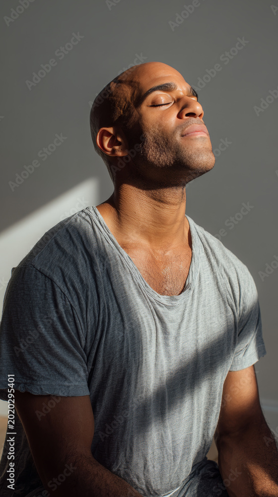 Obraz premium Serene young man with closed eyes enjoying peaceful sunlight indoors, wearing a casual grey t shirt. Calm expression and soft natural lighting create a tranquil, mindful atmosphere