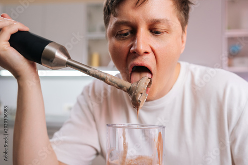 Billede på lærred Man tasting smoothie residue from an immersion blender blade with tongue, showing careless and risky behavior while preparing drink in modern kitchen
