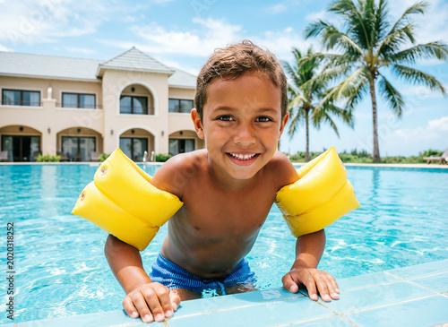 A child of European appearance, with a beautiful even tan, wearing inflatable armbands, holds onto the side of the pool, swims in the pool, looking at the camera, against the backdrop of a calf and pa