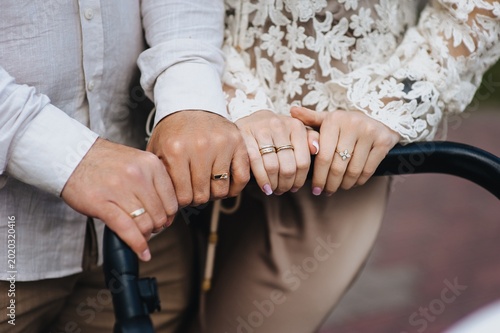 A close-up photograph of the hands of a young man, father, and mother holding onto a stroller with a newborn baby on a walk.