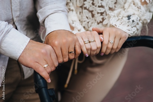 A close-up photograph of the hands of a young man, father, and mother of a woman, a family holding onto a stroller with a newborn baby on a walk.