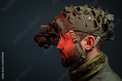 Tableau sur toile Side view profile of a bearded soldier wearing tactical helmet with night vision device and camouflage scrim on dark background with copy space
