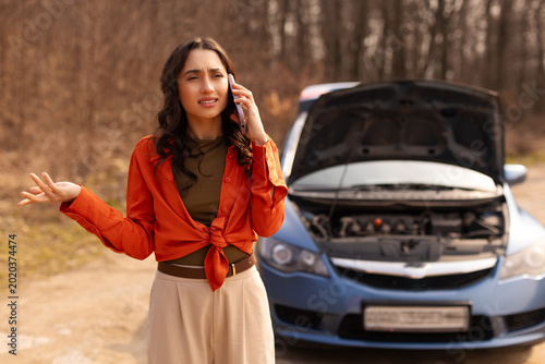 Fotografía Frustrated young woman talking on smartphone while standing beside broken down c
