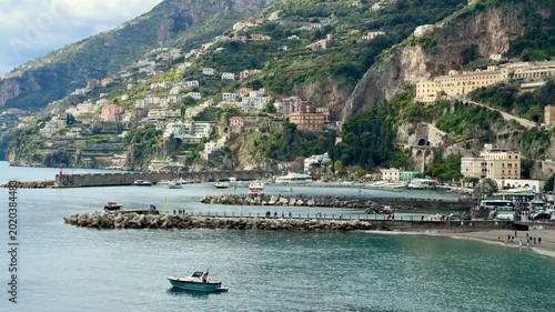 Scenic waterfront view of Amalfi Coast town in southern Italy with people walking along the seaside promenade, historic Mediterranean architecture, parked cars and dramatic mountain landscape