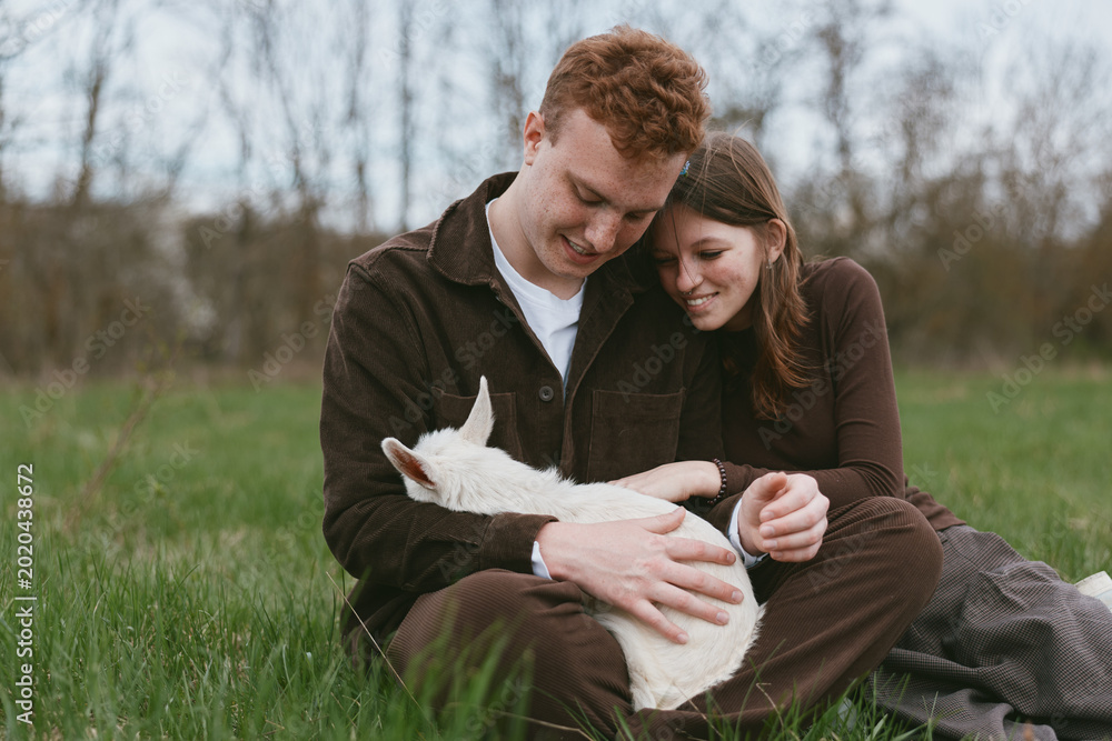 Obraz premium Loving couple sitting on a grassy field, gently holding a goat kid, enjoying outdoor life. Cozy, intimate moment capturing affection, nature, and simple happiness in a relaxed lifestyle scene.