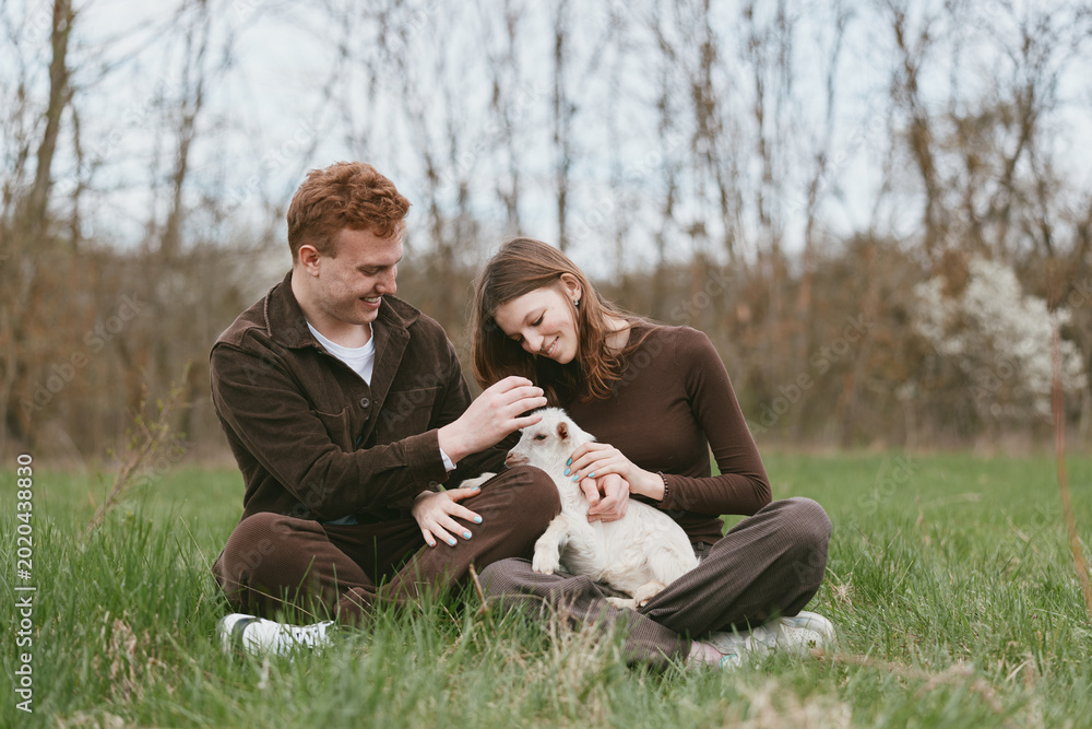 Obraz premium Cheerful couple outdoors with a friendly goat kid, sharing a calm bonding moment in a grassy field, natural light, casual outfits, warm atmosphere, gentle touch, countryside vibe, candid happiness