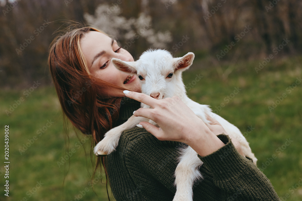 Obraz premium Smiling woman with long hair holding a white goat kid in a green field. Close-up on a natural backdrop. Lifestyle moment celebrating care, connection, and gentle affection between human and animal.