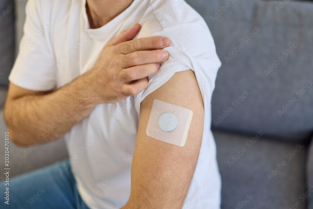 Fototapeta premium Man shows arm patch for diabetes sensor. The glucose monitor is a wearable medical device for health tracking at home, secured with adhesive on the arm. Healthcare monitoring and diabetic control.