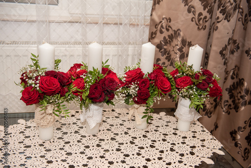 Decorative candles, suitable for weddings or baptisms, are decorated with red roses and white lace. The candles are adorned with red roses and white accents, symbolizing passion and elegance during sp