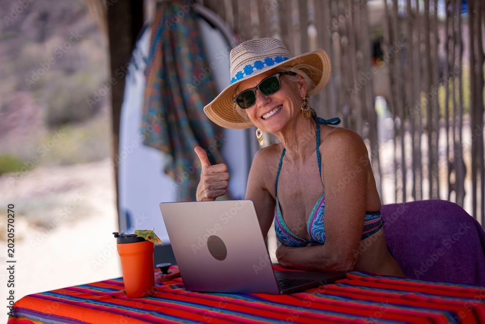 Fototapeta premium Mature woman works on a computer at a tropical beach, showing joy while enjoying her vacation in a sunny location