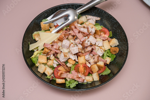 Caesar salad containing grilled chicken pieces, crispy bacon and cherry tomatoes. The salad is served in a modern black bowl and includes croutons and parmesan cheese shavings.