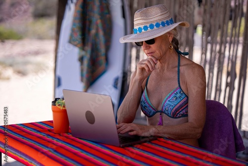 Konstfotografi A woman focuses on her laptop while seated at a beach table under a straw hat, e
