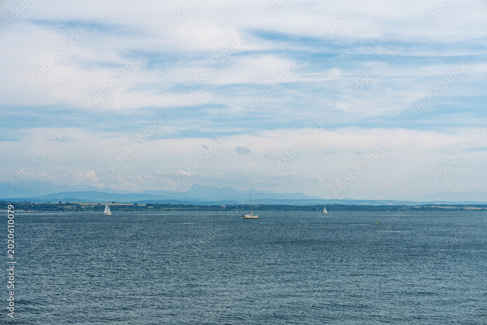 Obraz premium Sailboats glide across Lake Constance in Germany beneath a cloudy sky, framed by distant mountains and calm waters that convey a peaceful leisure scene and open natural vista.