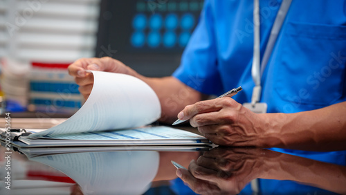 Quadro su tela Healthcare worker in blue scrubs reviewing paperwork on clipboard at hospital desk, holding pen, clinical setting with lab supplies and monitor in background
