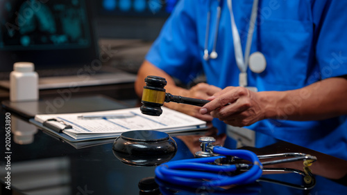 фотографія Close-up of medical professional in scrubs holding a judge gavel at desk with stethoscope, clipboard, and laptop, symbolizing healthcare law, malpractice, and regulation