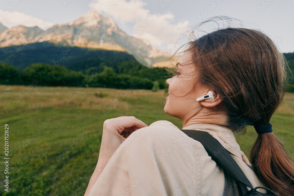 Obraz premium Woman enjoying outdoor landscape with headphones, hiking past mountains, casual backpack, serene mood, bright natural light, lifestyle moment in open field and meadow