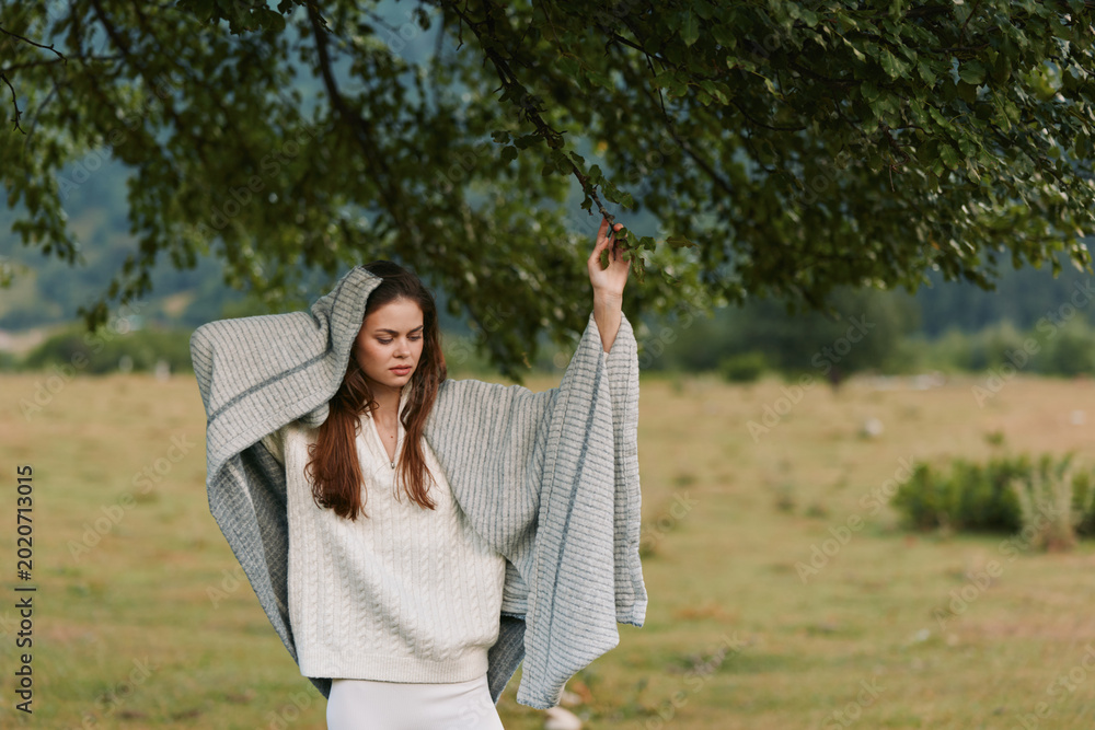 Obraz premium Cheerful woman outdoors, draped blanket and sweater, arms raised in a relaxed lifestyle moment, fresh air, soft breeze, natural colors, green meadow background in view