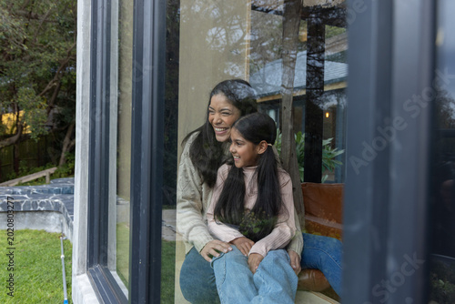 Indian mother and daughter sitting on cushioned seat, holding, looking through dark window at yard