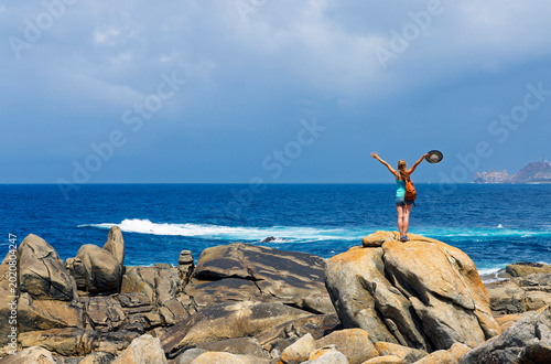 Foto Carefree woman standing on huge rock with arms raised holding hat looking at the Atlantic ocean in Galicia, Spain