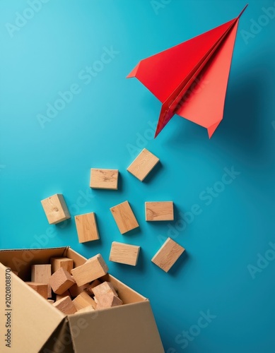 Red paper airplane flies over scattered wooden blocks spilling from a cardboard box. Concept of imagination  creativity  escape  and new beginnings.