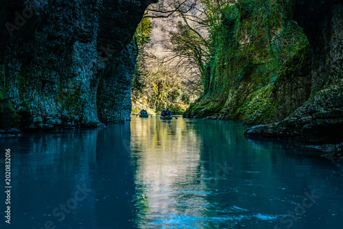 A small boat glides past a cascading waterfall in the turquoise waters of Martvili Canyon, with steep limestone walls and hanging greenery enclosing the narrow gorge.