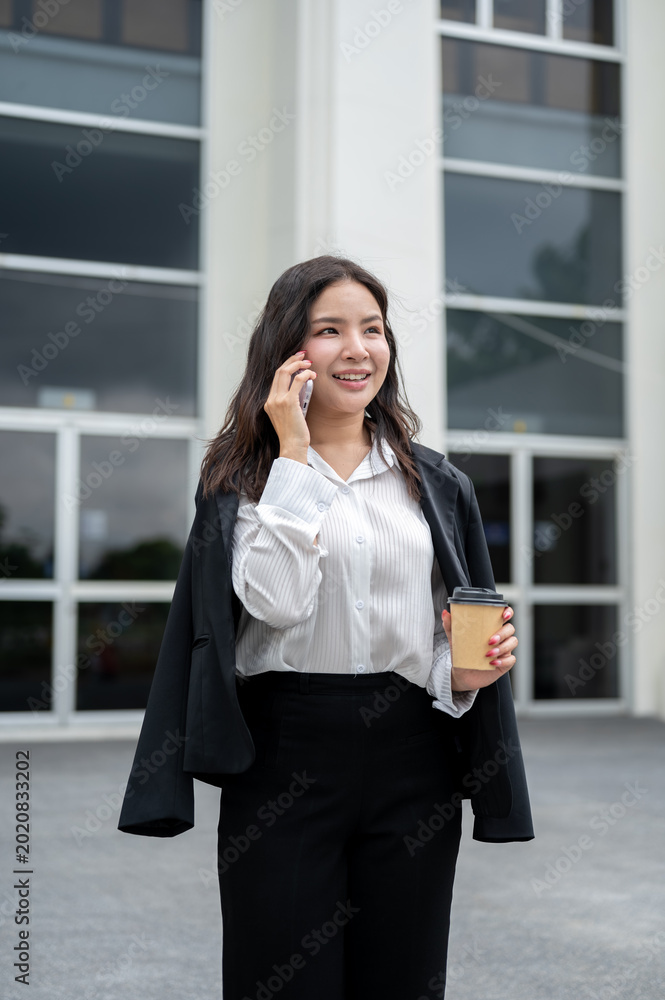 Fototapeta premium Pretty asian businesswoman holding a coffee cup talking on phone while standing outside the building