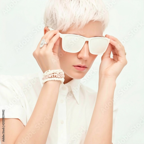 Fashion studio portrait of a woman with platinum blonde pixie hair wearing white sunglasses, a shirt, and beaded bracelets