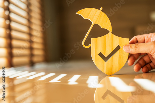 Hand holding wooden shield with checkmark and umbrella symbol on desk with sunlight. Life insurance, business protection, risk management, and reliable security guarantee concept.