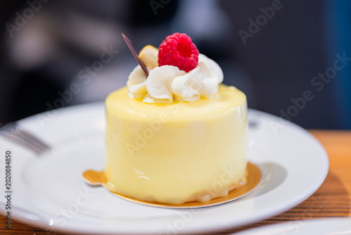 A close-up of a delicate lemon mousse cake or petit gateau with a smooth yellow mirror glaze. The dessert is topped with a swirl of white whipped cream and a fresh red raspberry, 