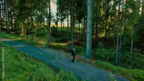 Male walking on forest trail in sunny weather. Explorer strolling along gravel path under clear blue sky. Person hiking through wooded area surrounded by trees and light