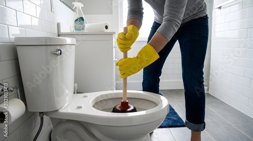 Person in yellow gloves plunging a clogged toilet in a clean, white tiled bathroom