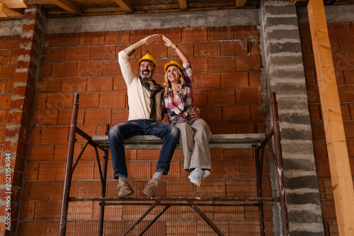 Two People Sit on a Scaffolding Inside a Building Under Construction in the Daytime While Wearing Yellow Hard Hats