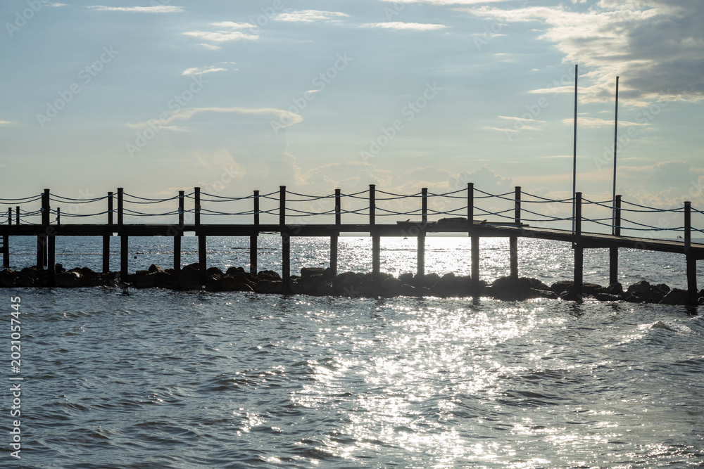 Fototapeta premium Wooden pier silhouette over shimmering sea water with bright sun trail under cloudy sky. Side, Turkey, Mediterranean.