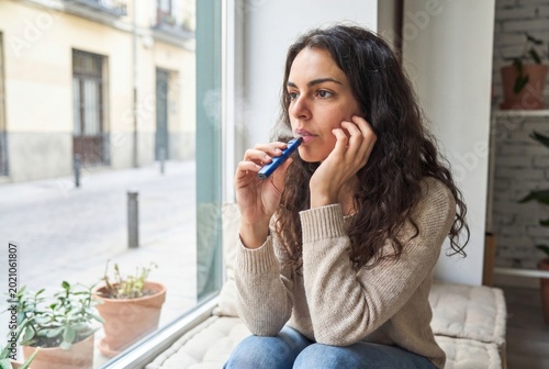 Young woman vaping electronic cigarette, looking thoughtful by a window overlooking a street