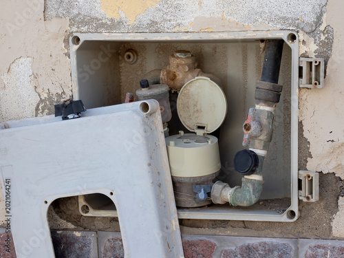 Open water meter box with plumbing pipes and damaged cover on exterior wall