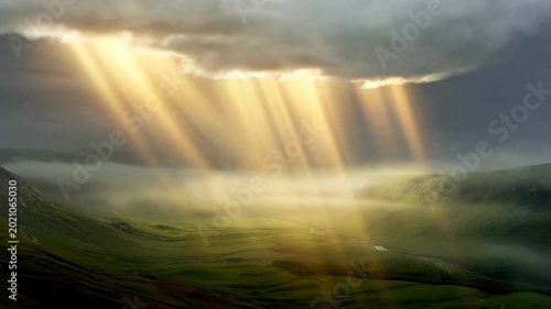 Time lapse aerial reveal of sunbeams piercing storm clouds and lighting green valley with misty hills at dawn in remote highland countryside