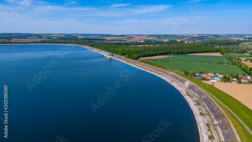 Aerial wide shot of Otmuchów Lake reservoir featuring the long concrete dam, hydroelectric power station structure, vibrant green forests, and rural landscape under a clear blue sky in bright daylight