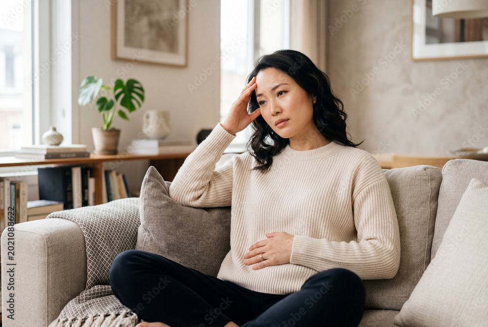 Obraz premium Sad asian woman sitting on sofa holding stomach and touching head