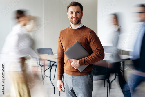 Confident businessman standing with laptop, smiling at camera as blurred colleagues walk past in a modern office environment